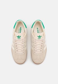 Beige suede sneakers with white stripes and green accents. Features a lace-up closure and textured rubber outsole.