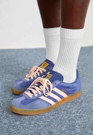 Personne portant des chaussettes blanches côtelées et des baskets Adidas Gazelle bleues avec des rayures rose clair et des semelles en gomme sur un sol recouvert de tapis bleu.
