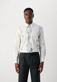 Λευκό πουκάμισο μακρυμάνικο με πράσινο αφηρημένο σχέδιο, με γιακά που κουμπώνει και υφασμάτινη υφή. Συνδυασμένο με σκούρες κομμένες παντελόνες.