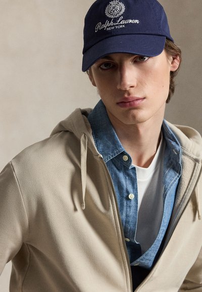Young man wearing navy Ralph Lauren cap, beige zip-up hoodie over a blue denim shirt and white t-shirt, neutral background.