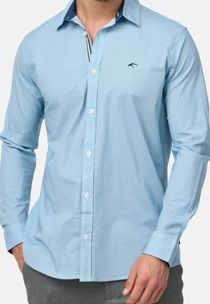 Homme portant une chemise à boutons bleu clair à motifs avec des manches longues et un pantalon gris, debout avec une main dans la poche.