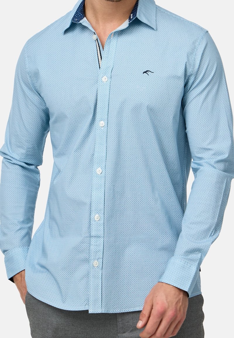 Homme portant une chemise à boutons bleu clair à motifs avec des manches longues et un pantalon gris, debout avec une main dans la poche.