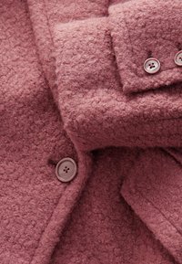 Manteau en laine rose texturée avec boutons arrondis, présentant une surface douce, un col chevauchant et des détails de couture visibles.