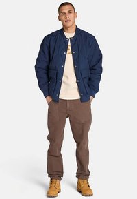 Marineblauwe doorgestikte bomberjack met drukknopen aan de voorkant, twee zakken aan de voorkant, gedragen over een crèmekleurige trui, gecombineerd met een bruine broek en gele laarzen.