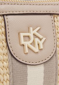 Bolso de cuero texturizado beige con acentos trenzados, presentando un parche marrón redondo y detalle del logo dorado. Patrón a rayas en tonos neutros.