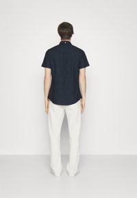 Koszula z krótkim rękawem w kolorze granatowym, wykonana z bawełny, z kołnierzykiem zapinanym na guziki i zaokrąglonym dołem, połączona z jasnymi spodniami i białymi butami.