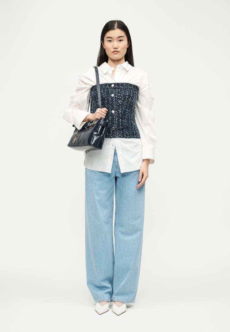 Femme portant une chemise blanche oversize avec un corset texturé bleu marine, un jean large bleu clair, des chaussures blanches pointues, et tenant un sac à bandoulière noir.