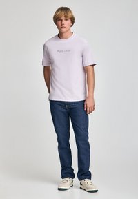 Jasnofioletowy t-shirt z nadrukiem "Polo Club", zestawiony z ciemnoniebieskimi dżinsami i białymi trampkami. Luźny krój i krótkie rękawy.