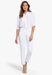 Witte geplooide blouse met knopen aan de voorkant, gecombineerd met witte rechte jeans. Het model draagt metalen sandalen, wat een schone, minimalistische outfit laat zien.