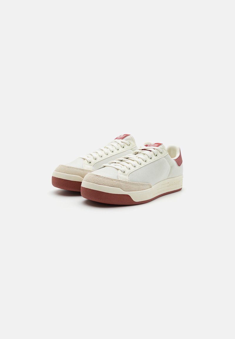 Baskets blanches avec une tige en maille texturée, accents rouges au niveau du talon et de la languette, et une coque en suède beige à l'avant. Semelle en caoutchouc rouge.