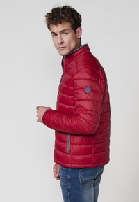 Rød polstret jakke med høj krave, lynlåslukning foran og sidelommer. Har et rundt logo-patch på venstre ærme og vandret syninger.