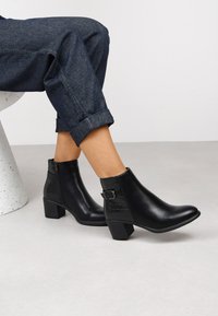 Stivali neri in pelle fino alla caviglia con tacco a blocco, chiusura con zip e fibbia decorativa. Dettagli con motivo coccodrillo sui lati. Indossati con denim arrotolato.