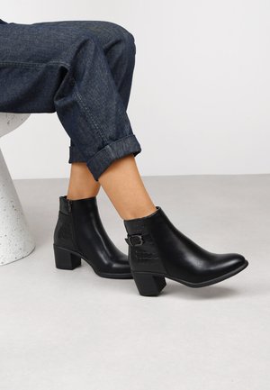 Bottines en cuir noir avec un talon bloc, fermeture éclair et boucle décorative. Accents en motif crocodile sur le côté. Portées avec un jean retroussé.