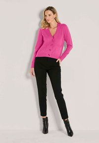 Frau trägt eine leuchtend pinke, knöpfbare Strickjacke, schwarze enge Hosen und schwarze Knöchelstiefel, steht vor einem schlichten, hellen Hintergrund.