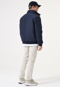 Marineblaue isolierte Jacke mit hohem Kragen, elastischen Bündchen und Reißverschluss vorne. Getragen über einem weißen T-Shirt und kombiniert mit beigen Hosen und grauen Sneakern.