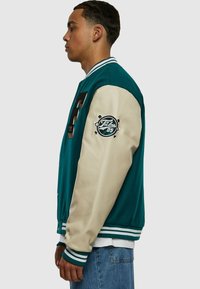 Jacheta varsity teal și crem cu patch cu litera "F", guler ribbed și manșete cu dungi. Prezentă un emblema pe mâneca stângă.
