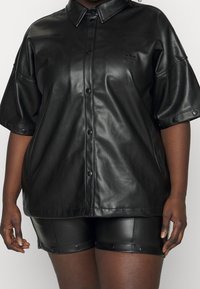 Svart, glänsande konstläder skjorta med korta ärmar, tryckknappar fram och en subtil logotyp på bröstet. Bärs med matchande shorts.