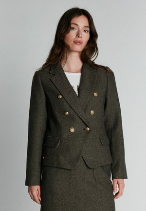 Femme aux longs cheveux bruns portant un blazer vert foncé à fines rayures avec double boutonnage, des boutons dorés et une jupe assortie sur un fond uni.