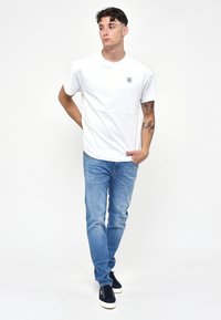 T-shirt di cotone bianco con un piccolo logo sul lato sinistro del petto, abbinata a jeans denim azzurri e sneaker scure, in piedi contro uno sfondo bianco.