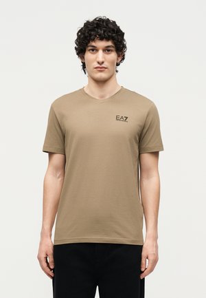 Jonge man met krullend donker haar, gekleed in een beige EA7 Emporio Armani t-shirt en zwarte broek, staand tegen een effen lichte achtergrond.