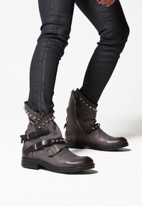 Brown leather ankle boots with a textured finish, metal studs, and black straps. Features a low block heel and a zip closure at the back.