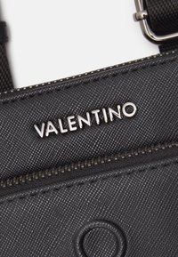 Detailní záběr na černou texturovanou kabelku Valentino se stříbrným zipem a kovovou sponou na popruhu.