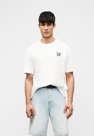 Jeune homme portant un t-shirt blanc avec un petit logo d'oiseau sur la poitrine et un short en jean bleu clair, debout devant un fond uni.