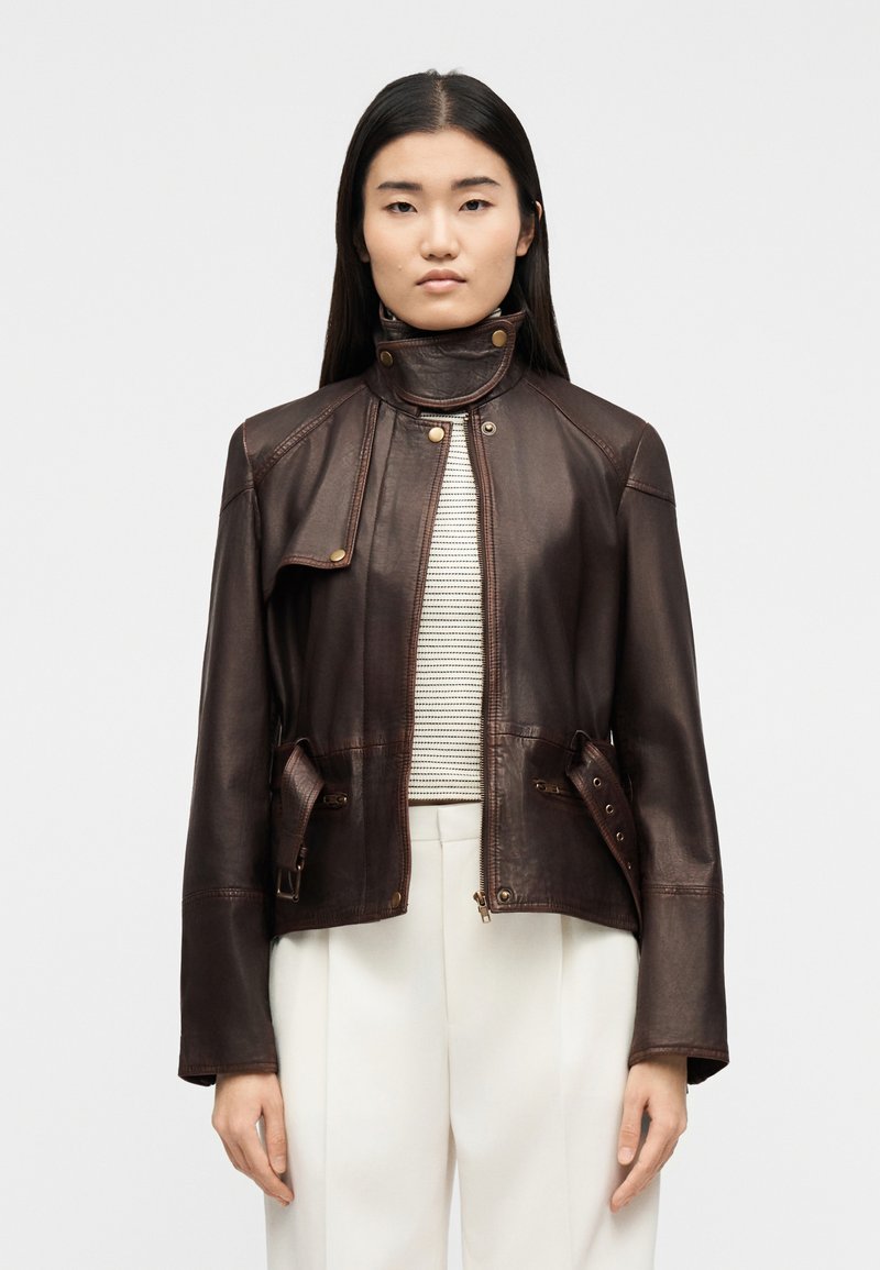 Γυναίκα με ίσια σκούρα μαλλιά, φορούν σκούρο καφέ δερμάτινο μπουφάν πάνω από μια ριγέ μπλούζα και άσπρο παντελόνι, στέκεται μπροστά σε ένα απλό φόντο.