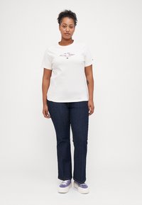 T-shirt en coton blanc avec le texte "Tommy Hilfiger", associé à un jean en denim foncé. Le modèle porte des baskets violettes et blanches. Design simple avec une coupe décontractée.