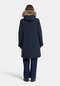 Laivastonsininen pitkä takki irrotettavalla karvavuorella varustetulla hupulla, jossa on sileä pinta ja normaali istuvuus, yhdistettynä leveälahkeisiin tummiin denim-housuihin.