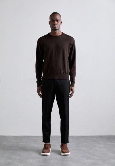 Hnědý pletený svetr, černé kalhoty a hnědé tenisky s bílou podrážkou. Oblečení má uvolněný střih a minimalistické designové prvky.