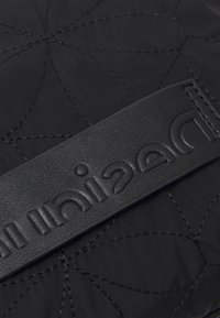 Schwarze gequiltete Tasche mit einem glatten schwarzen Lederträger, der geprägten Text, ein filigranes Stichmuster und eine strapazierfähige Stoffoberfläche aufweist.