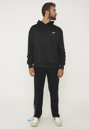 Uomo con felpa nera con cappuccio e pantaloni con striscia bianca, in piedi e che guarda alla sua sinistra, indossa scarpe da ginnastica bianche con etichetta blu.