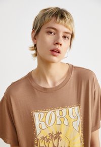 Bruine katoenen t-shirt met een grafisch ontwerp waarin "ROXY" in grote letters en palmbomen staat afgebeeld, korte mouwen en een ontspannen pasvorm.