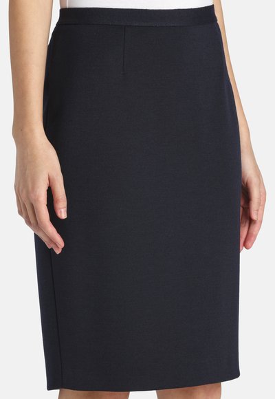 Kobieta ubrana w dopasowaną, czarną spódnicę do kolan o prostym kroju i wysokim pasie, ręce spoczywają wzdłuż ciała.