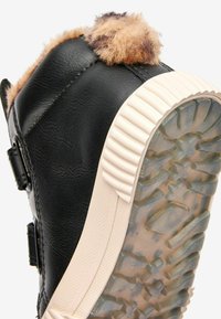 Svart vinterstövel med tjock beige gummisula och brun fuskpäls vid vristerna, sedd bakifrån och underifrån.