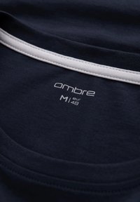 T-paita tummansinisestä puuvillasta, jossa on pyöreä kaula-aukko ja valkoinen ompelu, jossa on teksti "ombre" ja koko "M" valkoisella painatuksella.