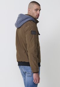 Braune gepolsterte Jacke mit grauem Kapuzenrand, ausgestattet mit einem schwarzen Patch am linken Ärmel sowie gerippten Bündchen und Saum. Getragen mit blauen Jeans.