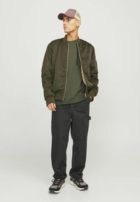Olivenfarbene Bomberjacke über einem dunkelgrünen Sweatshirt, kombiniert mit schwarzen Cargo-Hosen und grau-schwarzen Sportschuhen. Hellgraue Mütze.