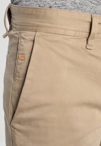 Beige bomullsbyxor med en slät textur, med en framficka och en orange etikett vid midjan. Sömmarna är minimala och diskreta.