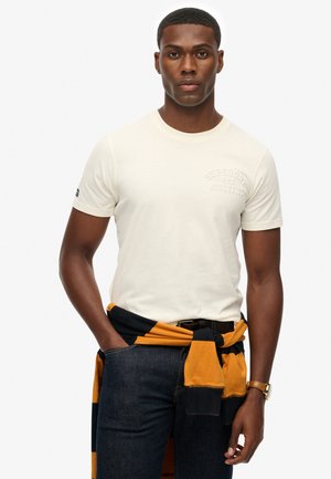 Light beige t-shirt with short sleeves and embossed "Superdry Athletics" logo. Worn with dark jeans, a black and gold striped sweater tied around the waist.