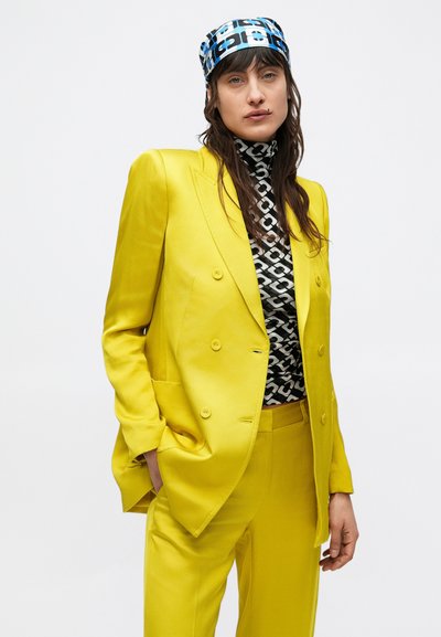 Blazer giallo doppiopetto con pantaloni coordinati, indossato sopra un dolcevita con motivo bianco e nero, completato da un foulard con fantasia.