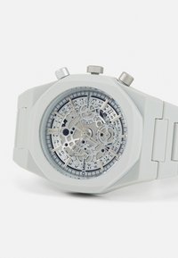 Montre octogonale blanche avec un cadran squelette transparent, accents argentés et bracelet en silicone. Possède des fonctions de chronographe et des marquages.