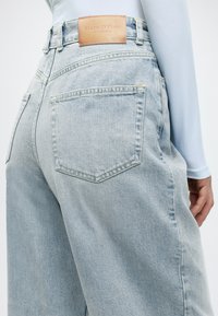 Hele sinised kõrge vöökohaga teksapüksid, millel on tagaküljel nahast silt, suured taskud ja kollased õmblused.