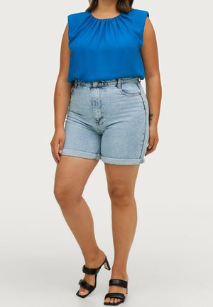 Woman wearing sleeveless royal blue blouse, light denim high-waist rolled shorts, and black heeled sandals, standing on plain background.