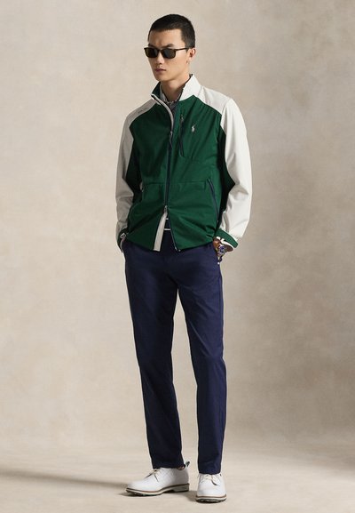 Homme portant des lunettes de soleil foncées, une veste zippée verte et blanche, un pantalon bleu marine et des chaussures blanches, debout les mains dans les poches devant un fond neutre.