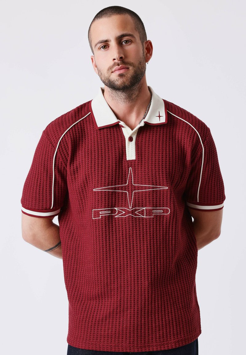 Homme aux cheveux courts et barbe portant un polo texturé bordeaux avec col blanc et logo "PXP", debout devant un fond clair uni.