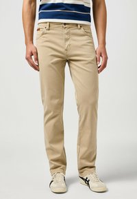 Pantaloni in cotone beige con vestibilità dritta, dotati di tasche frontali e una patta con logo, abbinati a sneakers chiare.
