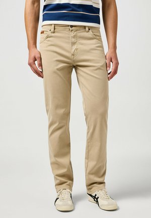Beige Baumwollhosen mit geradem Schnitt, ausgestattet mit Fronttaschen und einem Markenpatch, kombiniert mit hellen Sneakers.