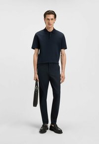 Marineblaues Poloshirt mit kurzen Ärmeln, taillierte marineblaue Hose, schwarze Slipper, hält eine schwarze strukturierte Tasche. Weicher Stoff, minimalistisches Design, taillierte Passform.